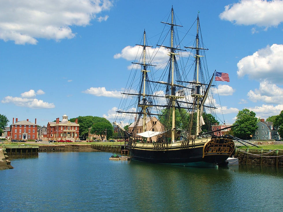 Historic tall ship docked, flying a U.S. flag, in a calm harbor. Red brick buildings and trees line the background under a blue sky.