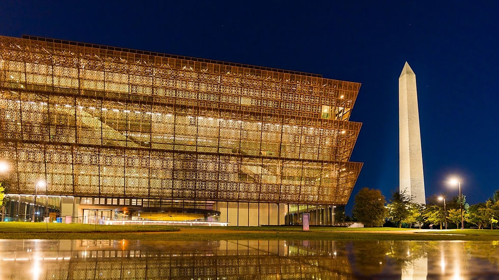 Lit museum with intricate design at night, reflecting in water. Washington Monument stands nearby against a deep blue sky.