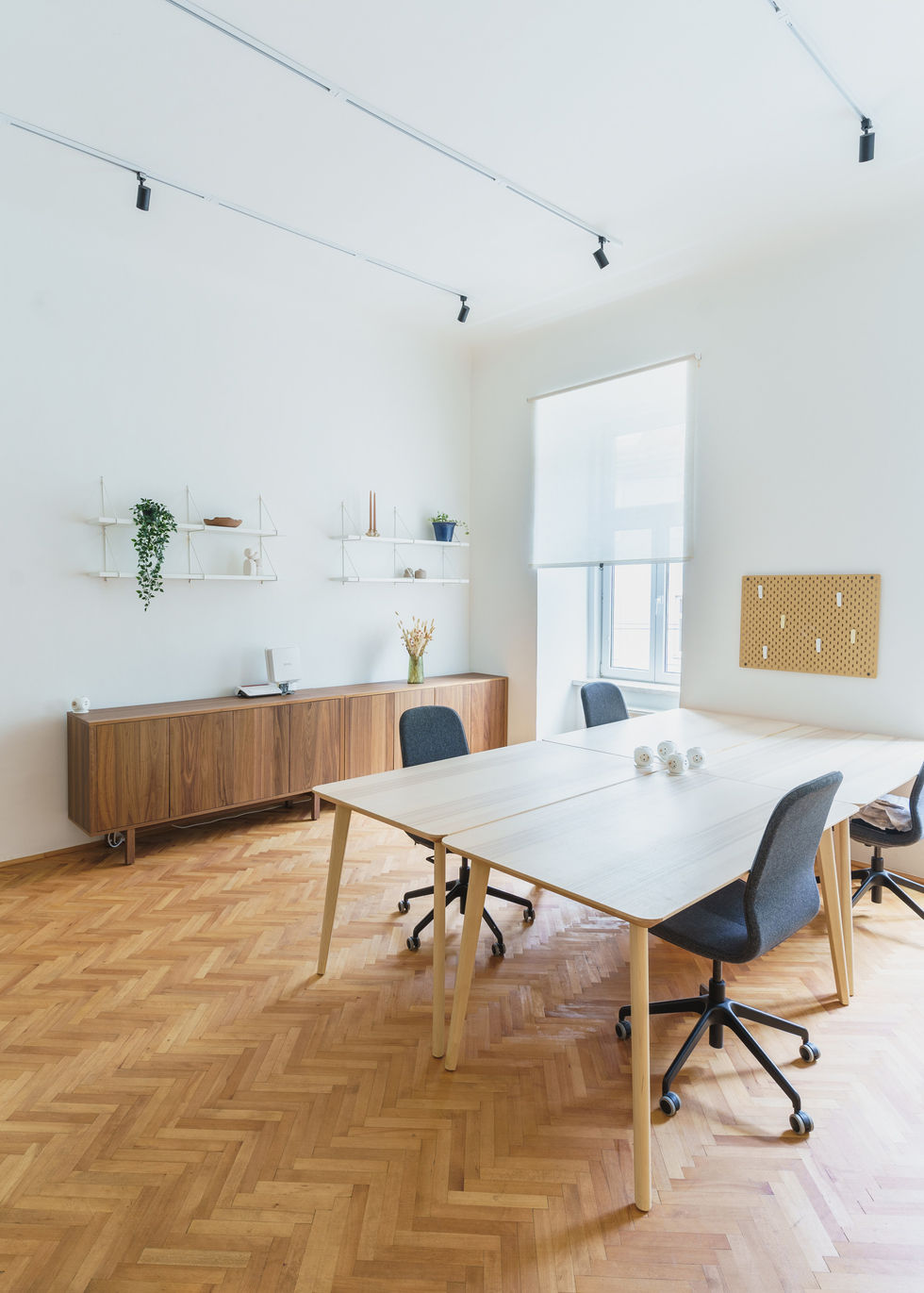 Ein Büroraum, sehr helle Lichtstimmung, das Tageslicht dringt durch die Fenster. In der Mitte Tische und Stühle, an der Seite eine braune Kommode.