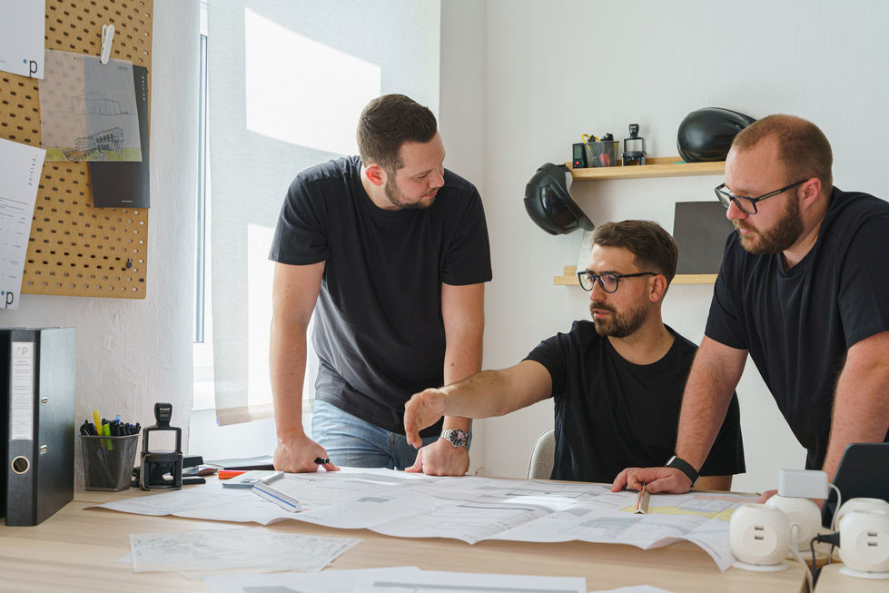 Drei Männer beraten an einem Tisch in einem Büro über ein Projekt, einer zeigt mit seiner Hand auf den Plan, welcher auf dem Tisch vor ihnen liegt.