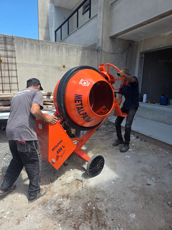Dois operários transportando uma Betoneira Metalpama 400L laranja da ALTMAXX em um canteiro de obras, demonstrando a mobilidade do equipamento para locação.