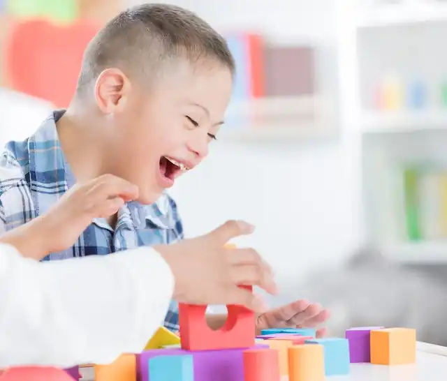 A smiling boy building with colorful blocks alongside his caregiver