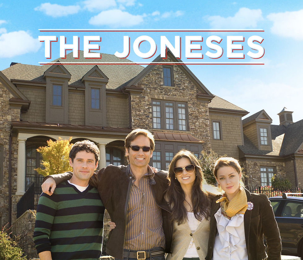 Four people smiling in front of a large stone house under a blue sky. "THE JONESES" text at the top. Casual attire, happy mood.