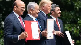 Four men in suits hold documents, smiling outdoors. Two folders are red and black. Green foliage in the background. Celebratory mood.