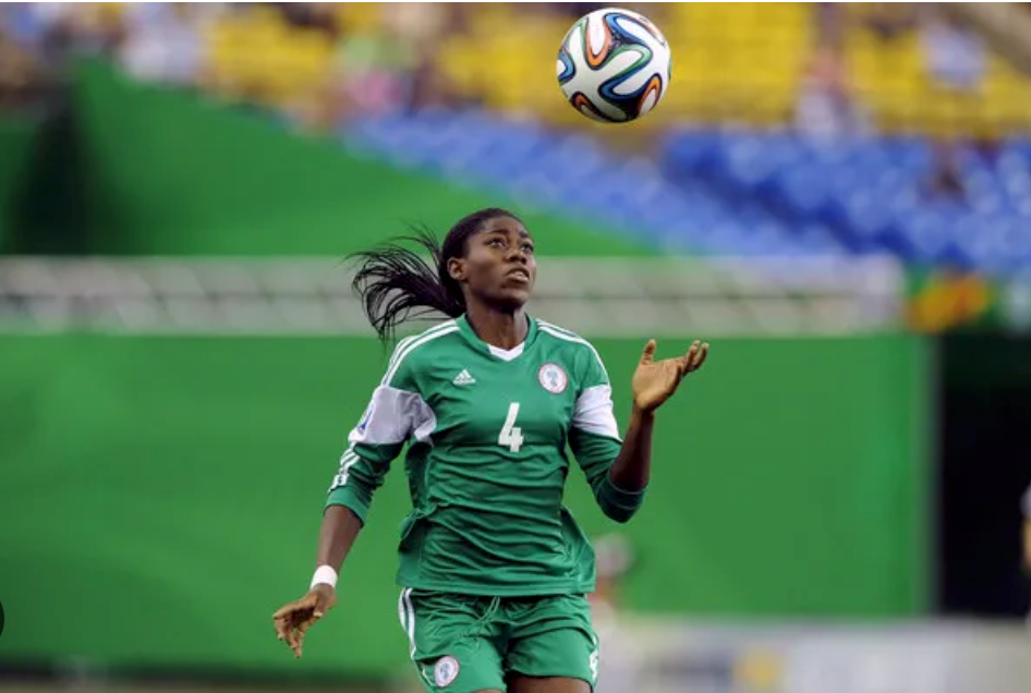 Female soccer player in green jersey #4 watches a ball overhead on the field, with blurred stadium seats in the background.