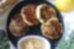 Four golden-brown fish cakes on a black plate with a creamy sauce, surrounded by kale, ginger, and a lemon on a light fabric.