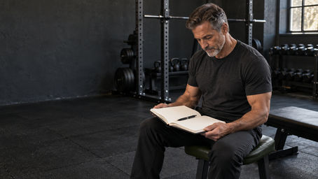 Mature man seated beside a gym bench reviewing an open training log, with a pen resting on the page.