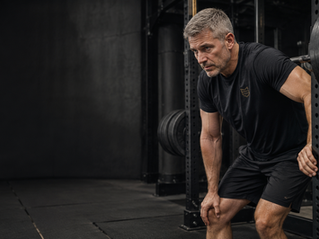 Middle-aged man leaning forward beside a loaded squat rack in an industrial gym, wearing a dark shirt with the MLS logo, with open shadowed space on the left side of the image.