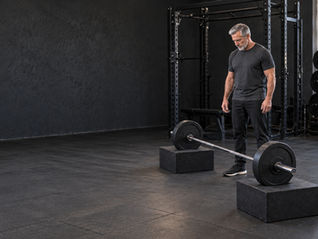 Mature man standing beside a lightly loaded barbell on pulling blocks in a quiet gym, looking down at the setup.