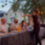 Server playfully pours liquid into a man's mouth at an outdoor dinner table. Evening lighting, trees, and houses in the background.