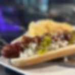 Close-up of a hot dog with ketchup, mustard, relish, and onions on a tray, against a blurred purple-lit background.