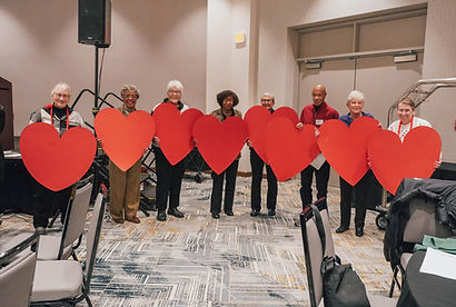 Seven smiling people standing in a row holding large red heart cutouts inside a conference room.
