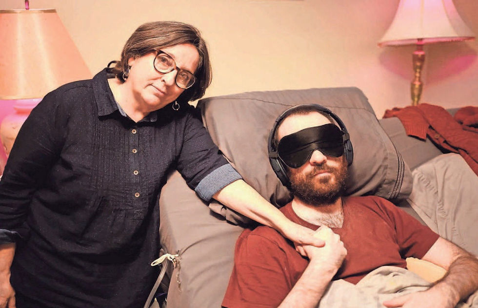 Galen Warden holding the hand of James Strazza, who is wearing noise cancelling headphones and a black sleep mask to protect him from the light.