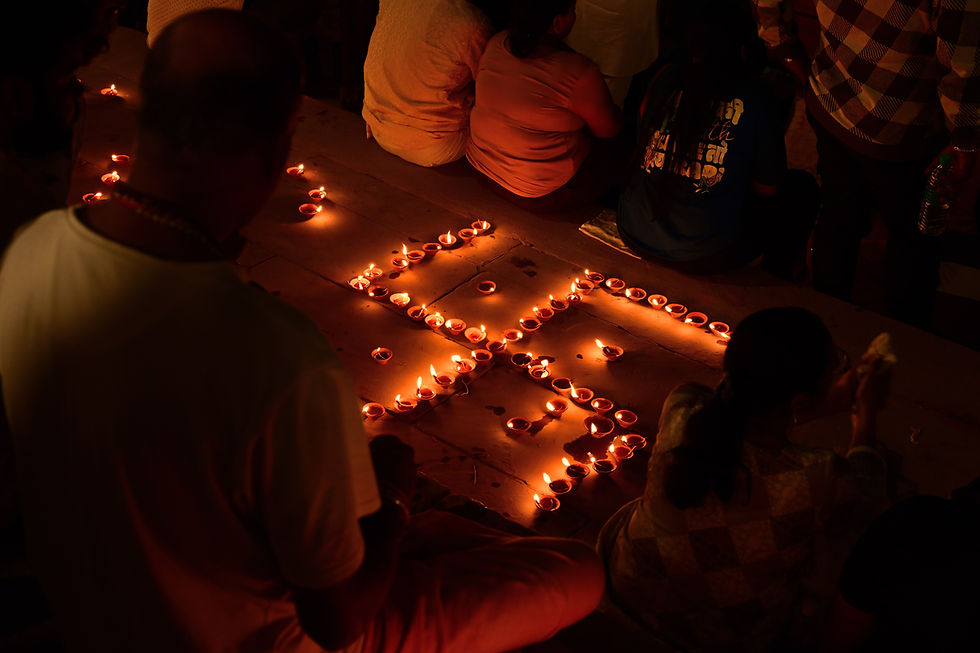 A close-up shot of small oil lamps (diyas) arranged on a dark surface to form a glowing swastika symbol, with the soft light illuminating the surrounding area where people are seated, at a Varanasi ghat during Dev Diwali.