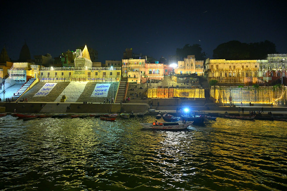 Jain Ghat in Varanasi illuminated at night with strings of golden and white ornamental lights draped over buildings and steps, and a large white structure on the ghat displaying the Hindi message 'अहिंसा परमो धर्म' (non-violence is the supreme virtue).