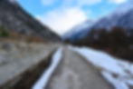 Snow-lined cement path flanked by stone boundary and pine-covered ridges under blue sky, Rakcham, Himachal Pradesh.