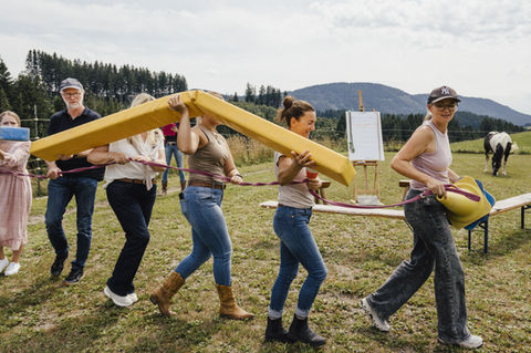 Ein Team bei einem Gruppen - Team Coaching Event / Team Event mit Pferden draußen auf einer Wiese. Im Hintergrund Flipchart und Pferde. Die Gruppe trägt an einem Seil verbunden Material zum Aufbau eines Parcous.