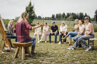 Jana Schöpper und Simone Krebs sitzen vor einer Gruppe. Szenerie ist draußen, grüne Wiese, Sommer. SaddleUp Coaching - Team Events.