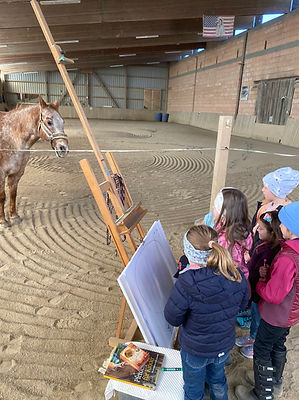 Viele junge Kinder stehen vor einer Malunterlage und wollen etwas einzeichnen. Im Hintergrund steht das Pferd und Co-Trainer und schaut interessiert zu.