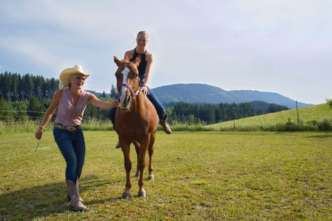 Jana sitzt auf Coaching Pferd und Simone mit Cowboy Hut hält das Pferd. Idyllisch auf der SaddleUp Location, grünes Allgäu mit Hügeln und Wald im Hintergrund.