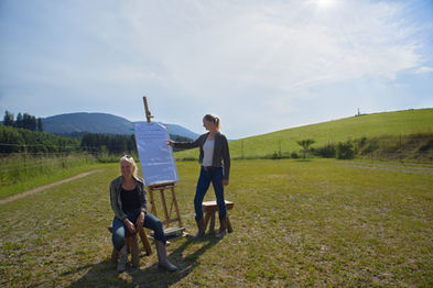 Jana Schöpper steht an einem Flip Chart, Simone Krebs sitzt davor. Hintergrund Wiese, Wald und Hügel. Outdoor Coaching im Allgäu mit Pferden.