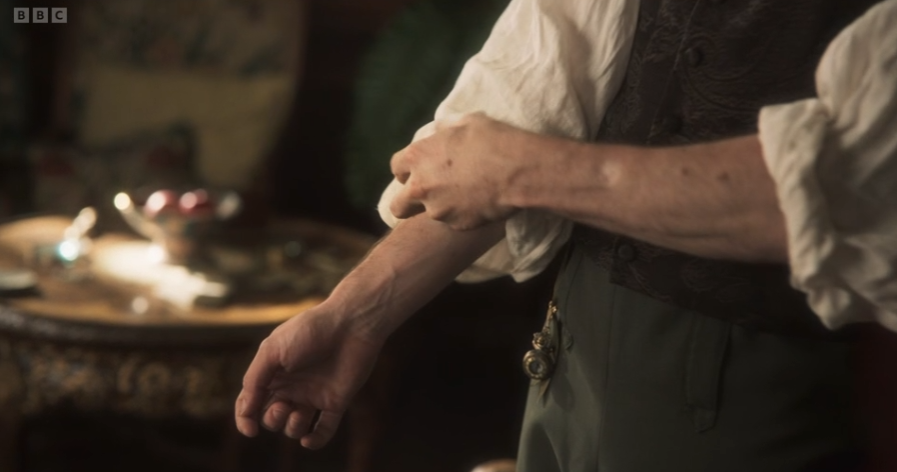 A person in a vest rolls up their sleeves, revealing forearms, in a warmly lit room. A wooden table with a bowl of apples is in the background.