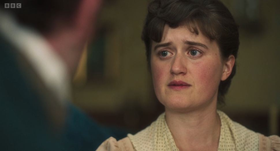 Worried woman in a cream blouse looks at a blurred man in the foreground, with a BBC logo in the corner.
