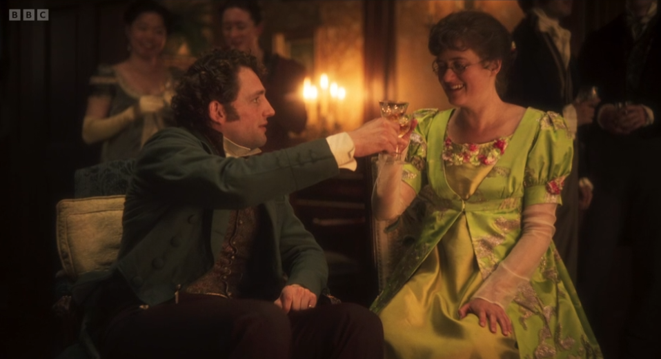 BBC logo. A man and woman in period dress smile and toast with glasses at a candlelit party, guests blurred in the background.