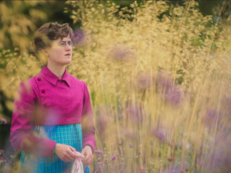 A young woman in a magenta jacket stands amongst wheat and flowers