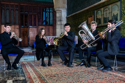 Five musicians in black suits play brass instruments in a richly decorated room with wooden panels and green patterned walls.