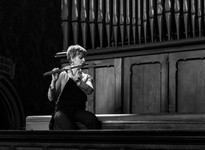 A woman plays the flute seated on a wooden bench in front of a large organ. The setting is dimly lit, creating a serene atmosphere.