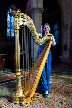 A woman in a blue dress smiles while holding a large ornate harp in a dimly lit church with stained glass windows.