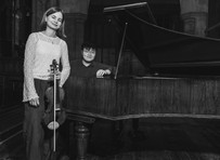 Woman holding a violin stands beside a grand piano with a seated person. Ornate room, monochrome, calm expressions.