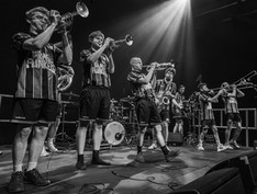 a line of musicians at front of stage playing brass instruments including trombones, trumpets and saxaphone