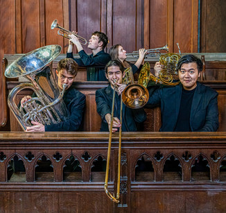 Five musicians in black attire play brass instruments in a wooden-paneled setting, creating a lively and focused atmosphere.