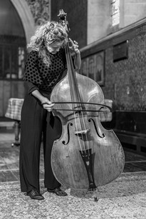 Musician with curly hair plays a double bass in a vintage room with patterned walls and arched window. Black and white image.