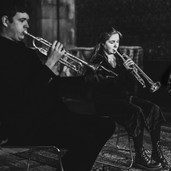 Two musicians play trumpets in a dimly lit room. The focus is on their instruments and expressions, creating an intense, artistic mood.