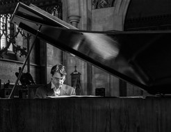 A man in a suit plays a grand piano in a dimly lit, ornate church. The mood is somber, with intricate stained glass in the background.