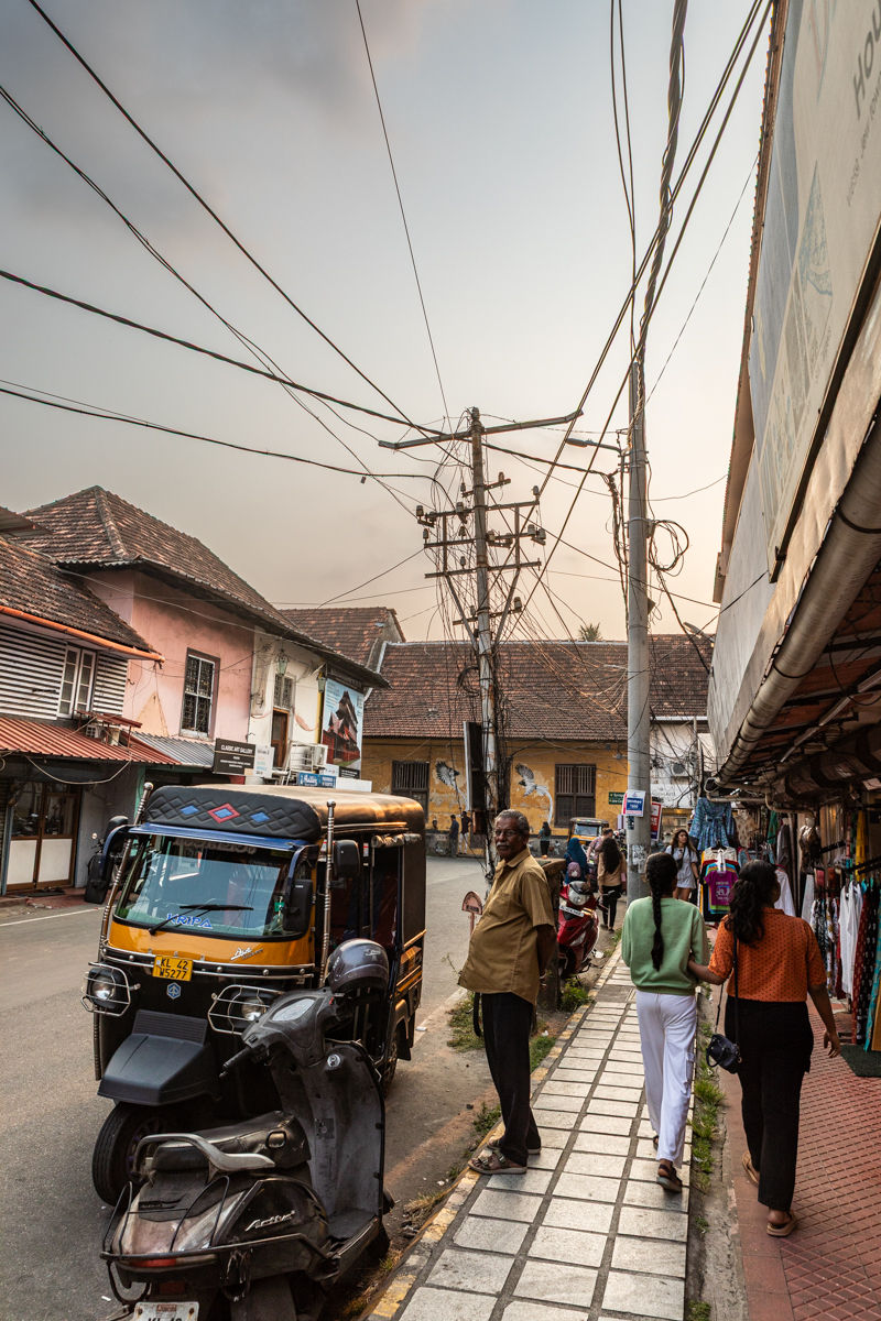 A busy street scene with a tuk-tuk, pedestrians walking by shops. Overhead wires form a web, creating an urban atmosphere. Mood is lively.
