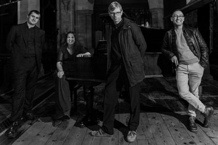Four people in a dimly-lit room with a grand piano. Three stand, one sits, smiling. Wooden floor, stone pillars. Black and white image.