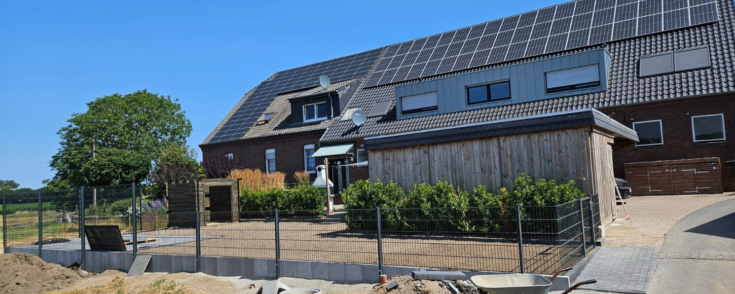 Ein anthrazitfarbener Doppelstabmattenzaun verläuft entlang einer Reihe von Beton-L-Steinen zur Hangsicherung vor einem großen Wohnhaus mit Solaranlage auf dem Dach und einer Holzverkleidung im Erdgeschoss.