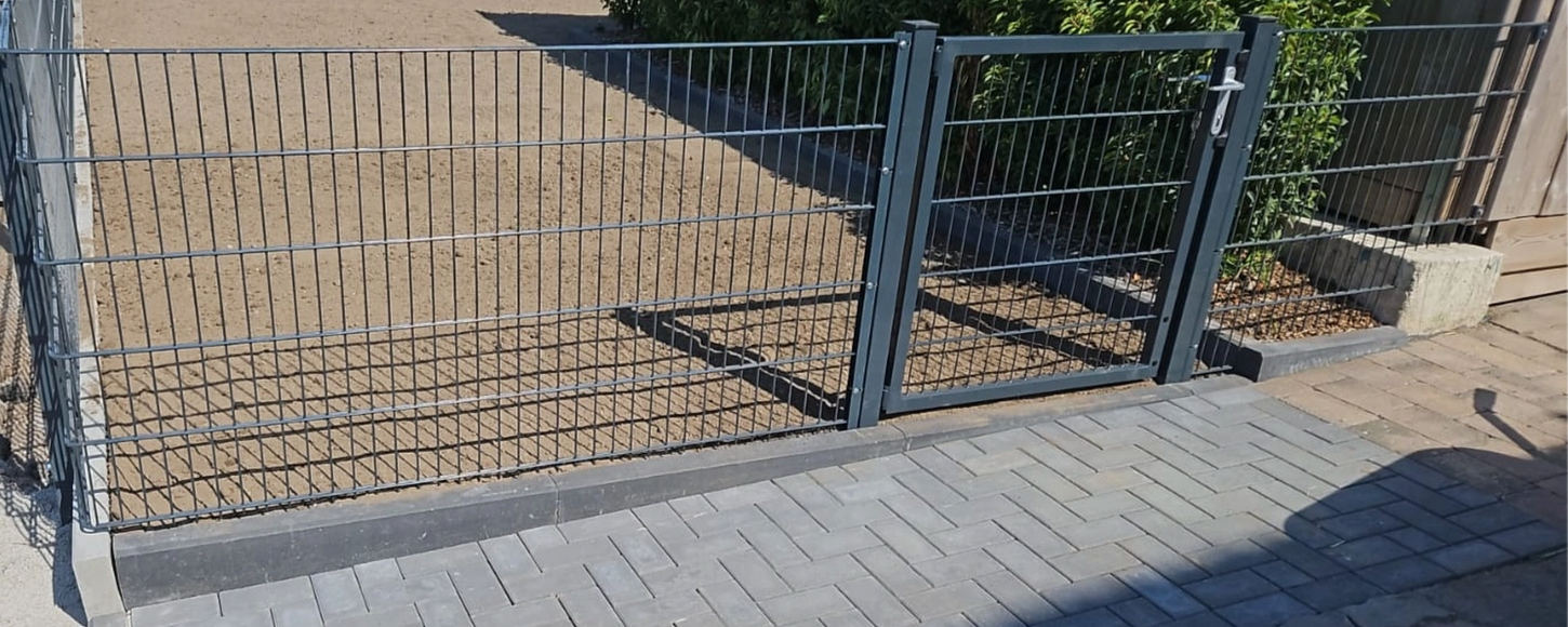 Modernes anthrazitfarbenes Gittertor mit Griffschale und Profilzylinder, integriert in einen Metallzaun auf L-Steinen, neben einem grauen Pflasterweg.