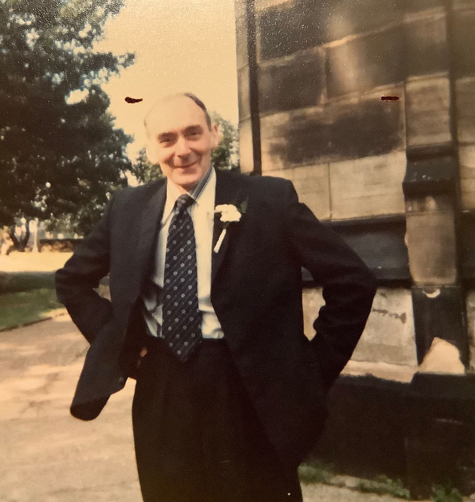 Smiling man in a suit with a flower boutonniere poses outside near a stone building. Trees in the sunny background create a warm ambiance.