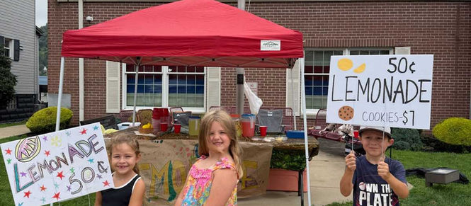Three children stand at a red-canopy lemonade stand outside a brick house, holding signs for 50¢ lemonade and $1 cookies.