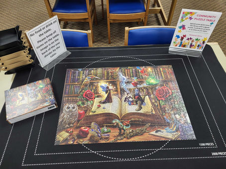 Library puzzle table with a completely finished fantasy jigsaw, signs reading no food or drink and community puzzle table, blue chairs nearby.