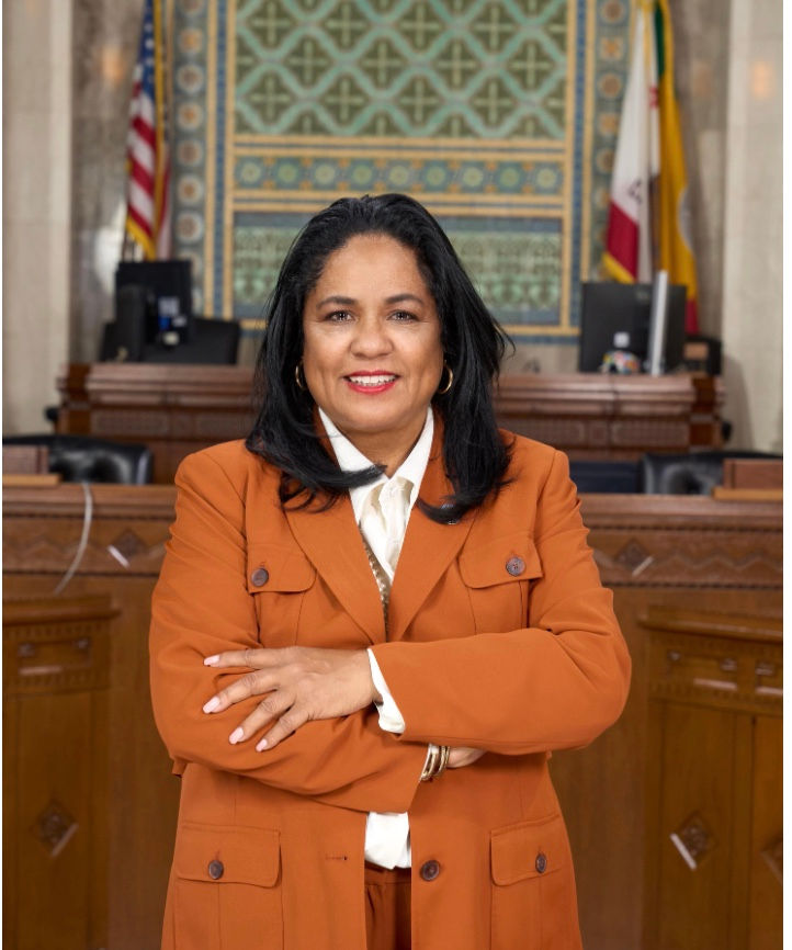 African American woman standing in city council chambers in front of CA flag and US flag. She is wearing white blouse with orange blazer.
