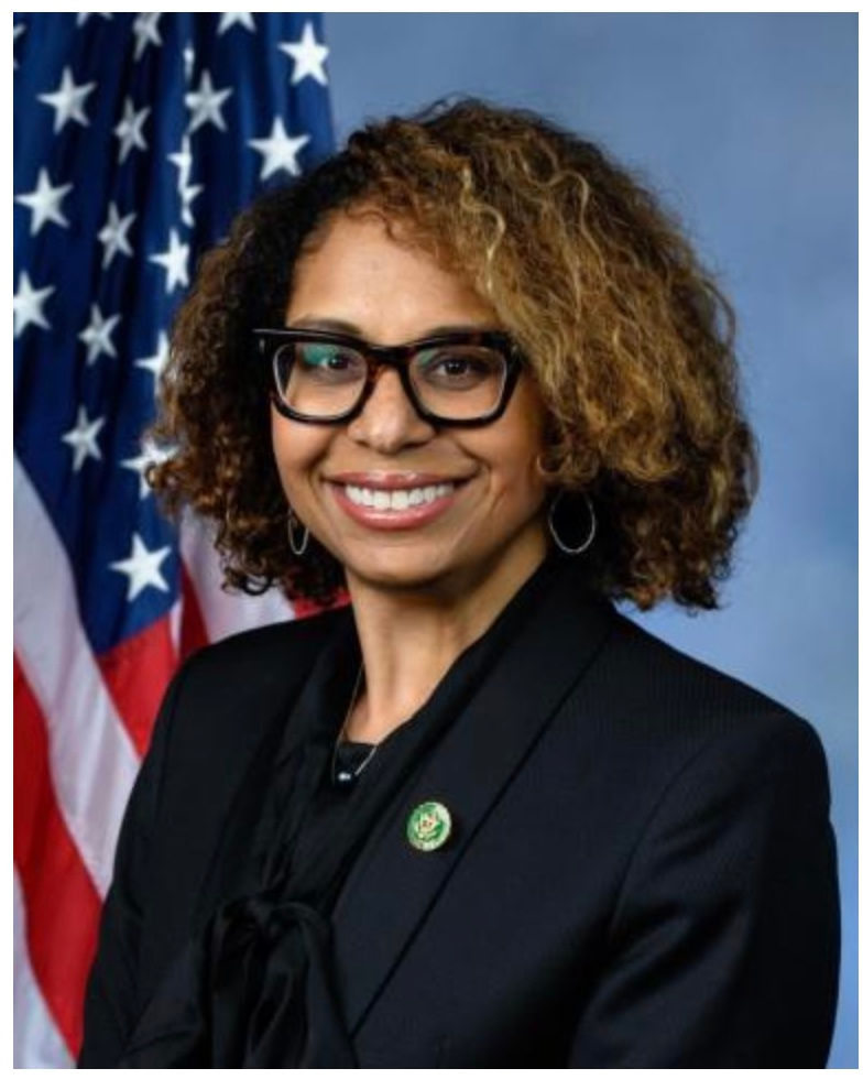 African American woman with curly hair and large framed glasses wearing black blazer and standing in front of US flag