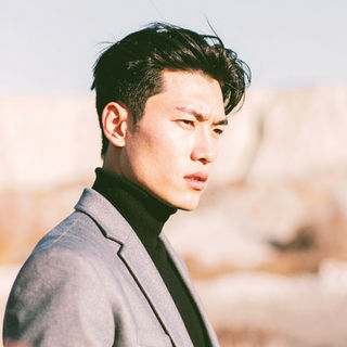 グレーのスーツと黒のタートルネックを着た日本人男性モデルが、冬枯れの野原で遠くを見つめる。映画のワンシーンのような構図と自然光が特徴のポートレート。