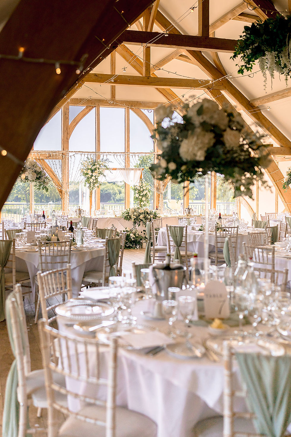 A room set with flowers and glasses for a wedding breakfast with sun coming through the windows behind.