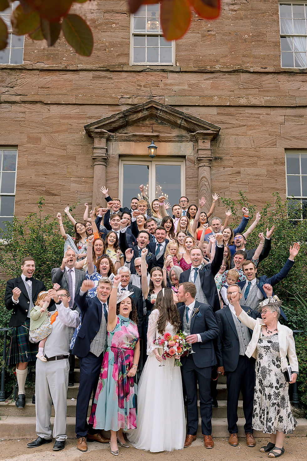 A family group shot outside Newton Hall at a wedding with the bride and groom kissing